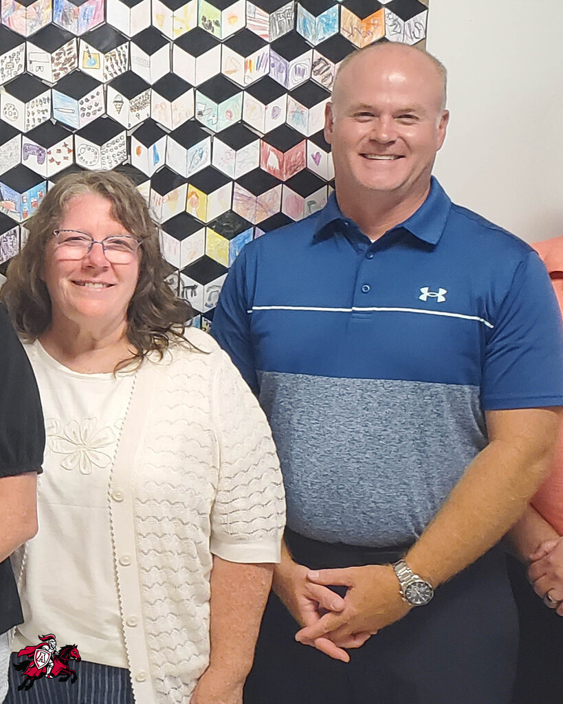 A man and a woman smile at the camera. The man wears a blue and gray polo, and the woman wears a white cardigan. Behind them is a colorful tile wall.