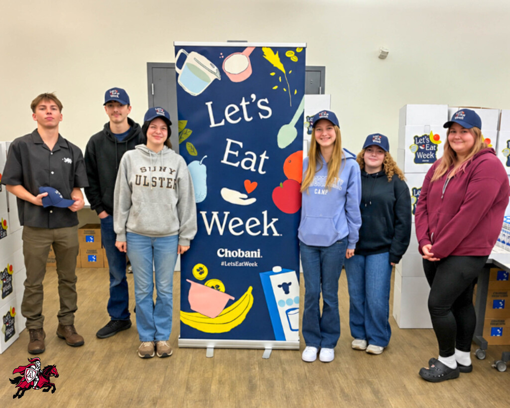 Six people stand behind a banner that reads, "Let's Eat Week." They wear hats and sneakers. A refrigerator is visible in the background.