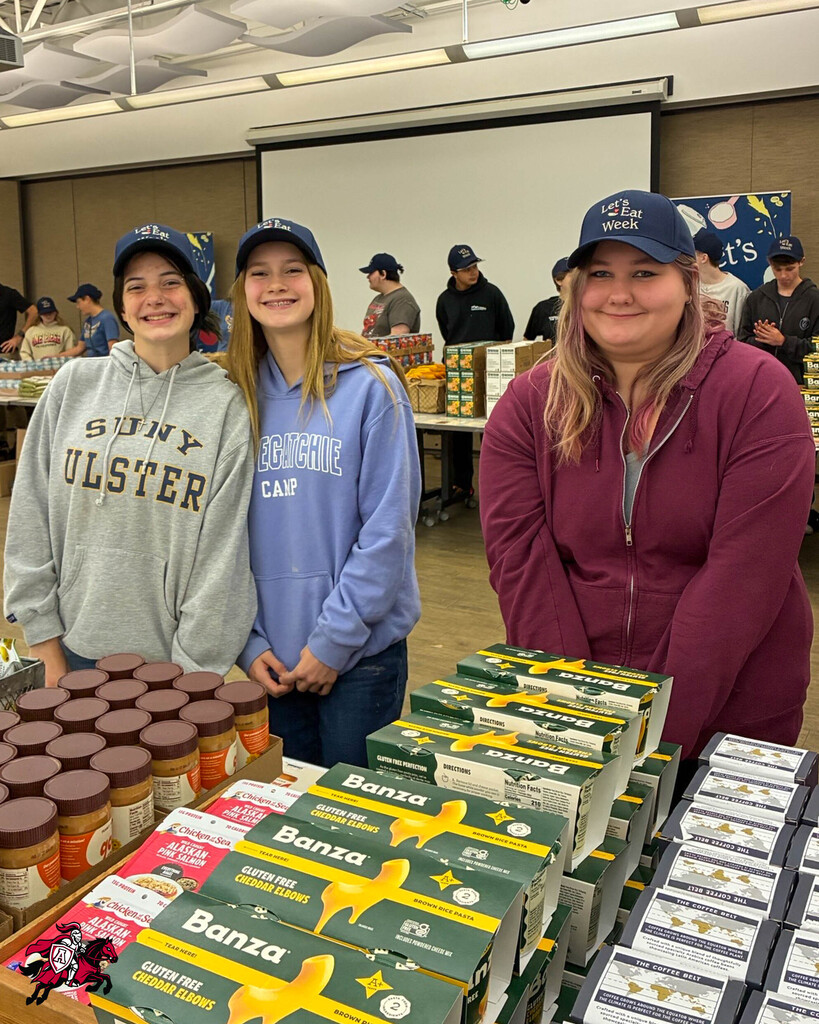 Three women, each wearing a cap, stand beside a table with jars of peanut butter and Banza boxes.