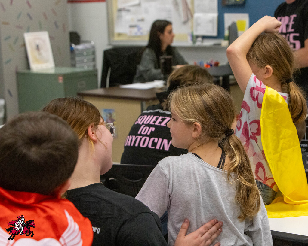 Kids seated in a classroom. One girl wears a yellow cape. A teacher works at a desk.