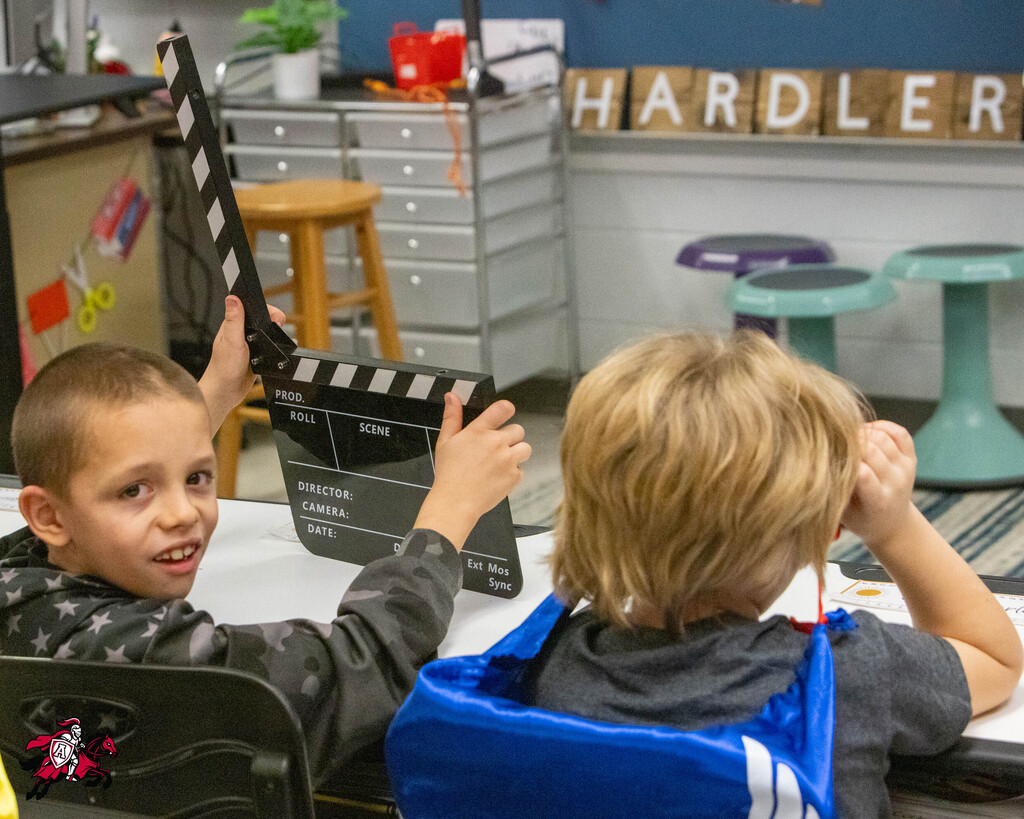 Two young children seated at a table, one holding a clapperboard, with classroom decor in the background.