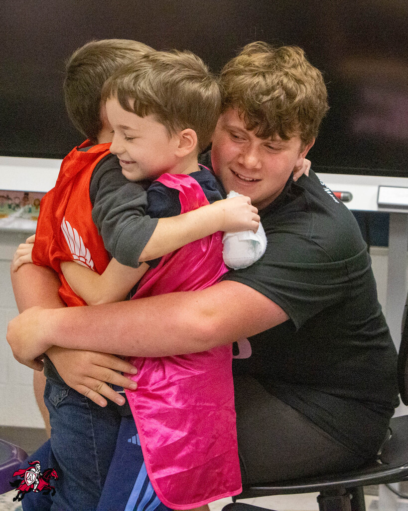 Three boys, one wearing a pink cape, hug each other in a classroom setting. One boy sits on a chair.
