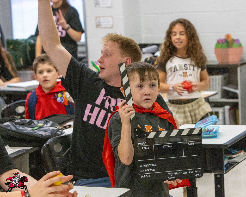 A young boy holds a clapperboard in front of a seated child. Behind them, a boy waves a red cape. A teacher sits next to them, and a girl stands behind them in a classroom.