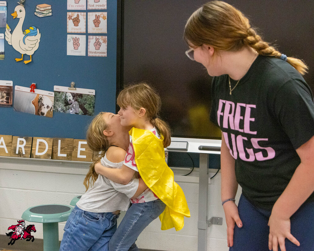 Two children hugging indoors, one holding a yellow cape. A woman with glasses watches. Background includes a bulletin board.