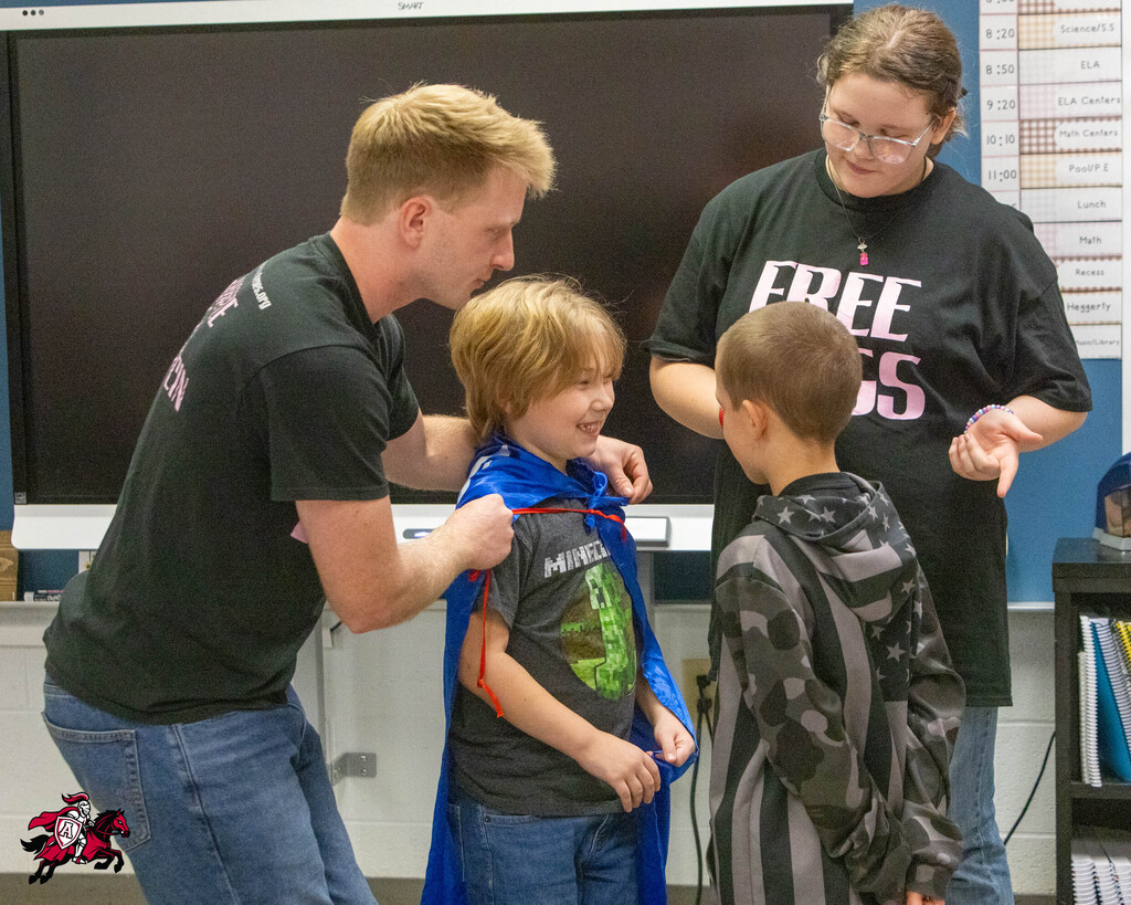 A man helps a boy put on a cape in a classroom. A woman stands next to them.