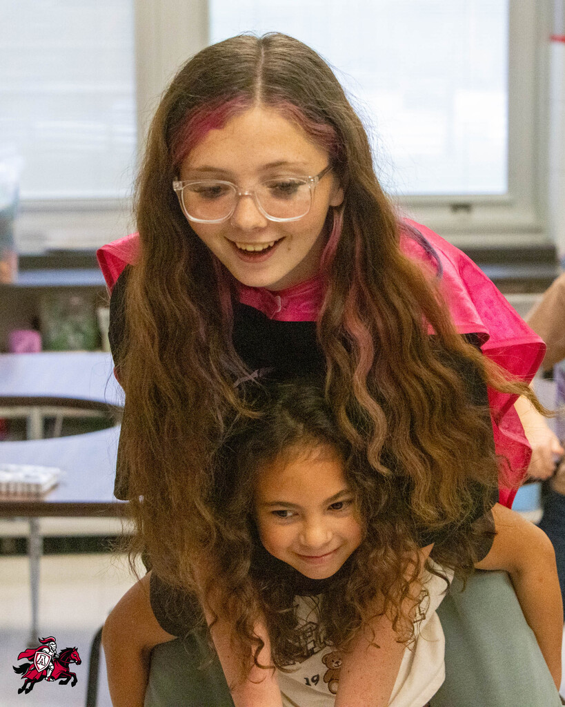 Two children in a classroom. A girl with long hair and glasses carries another girl on her shoulders.
