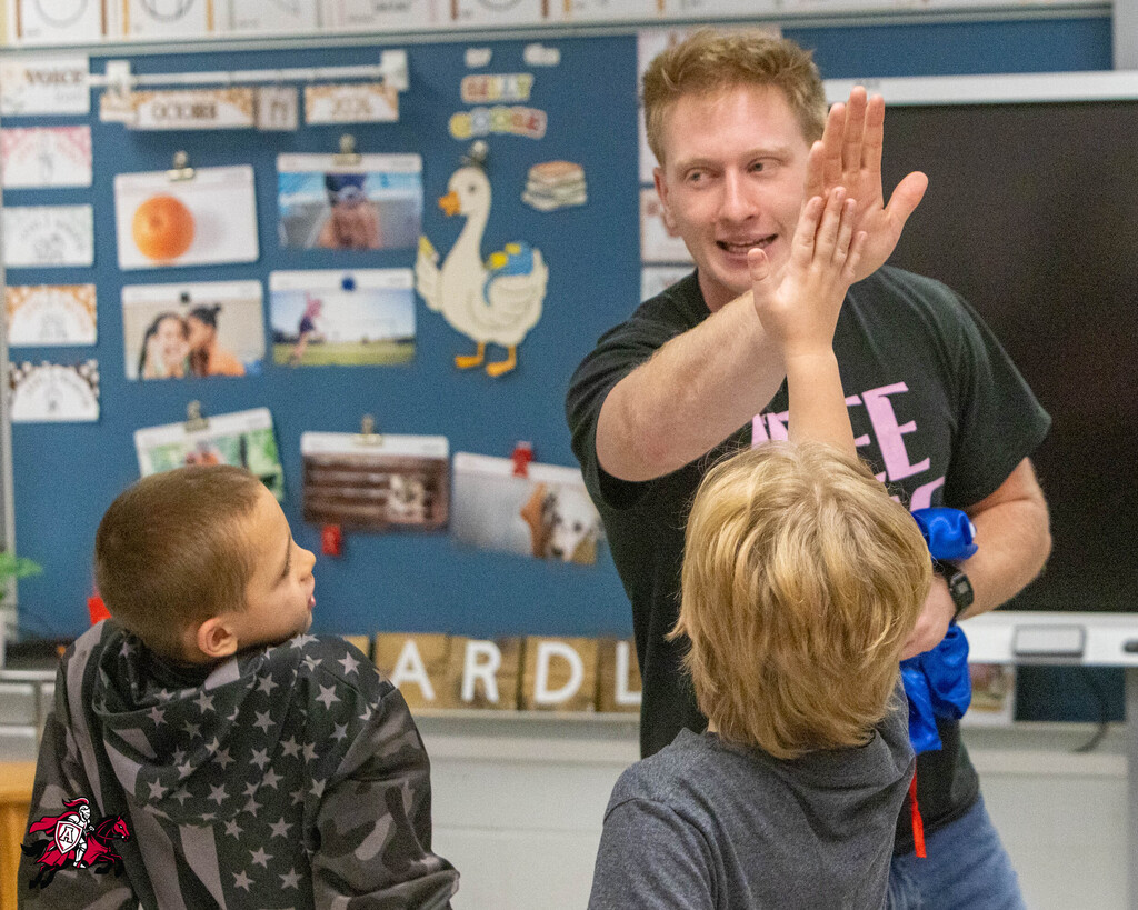 Man high-fiving two kids in a classroom with a bulletin board and a monitor in the background.