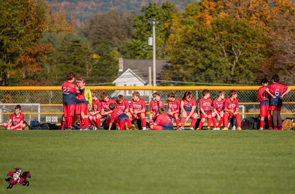 A team of players in red uniforms stands in a group on a soccer field. Trees and houses in the background.