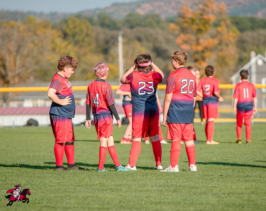 Youth soccer players in red and blue uniforms, some with numbers, standing in a group on a grassy field.