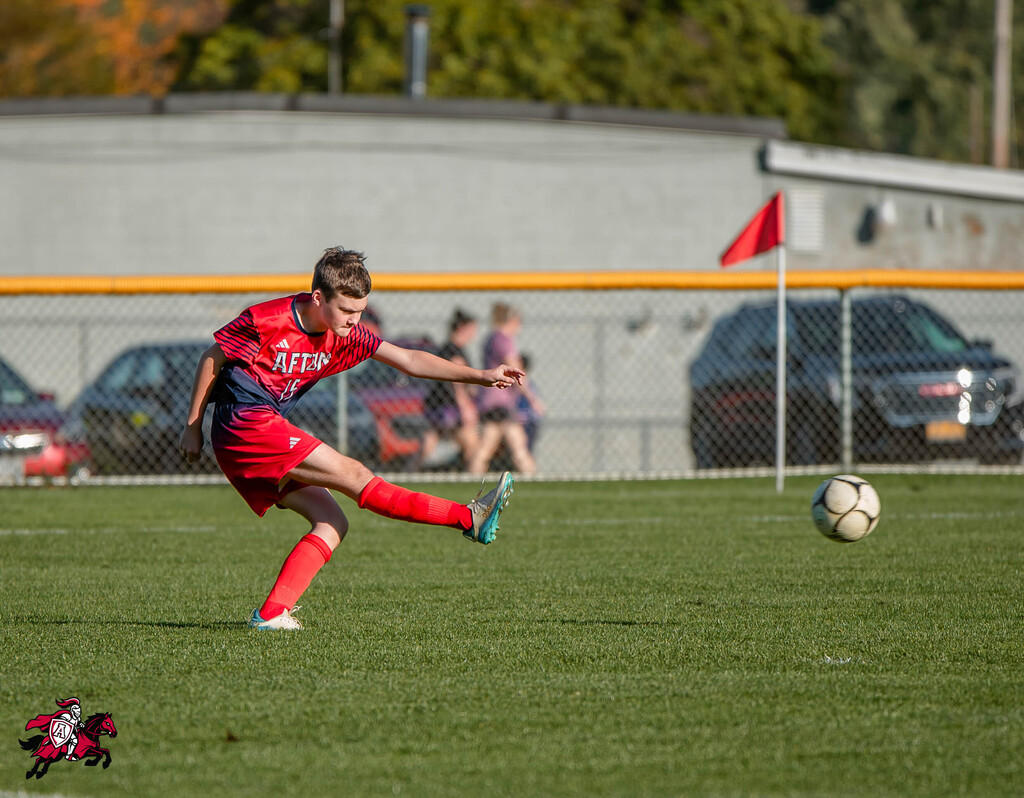 A soccer player in red uniform kicks a ball on a field with a fence and parked cars in the background.