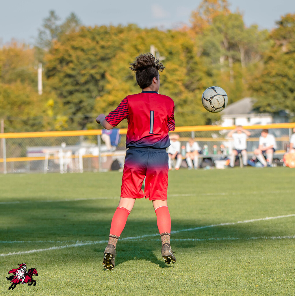 A player in red kicks a ball in a soccer game. Trees in the background.