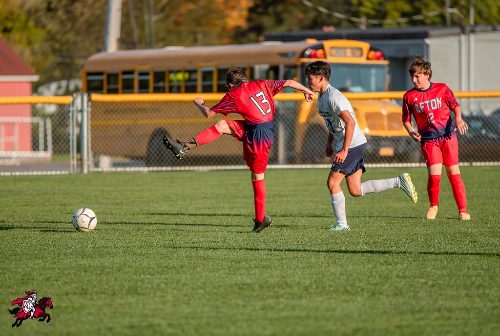 Three soccer players on a green field, one in red with number 13, another in white, and one in red with number 9. A yellow school bus is in the background.