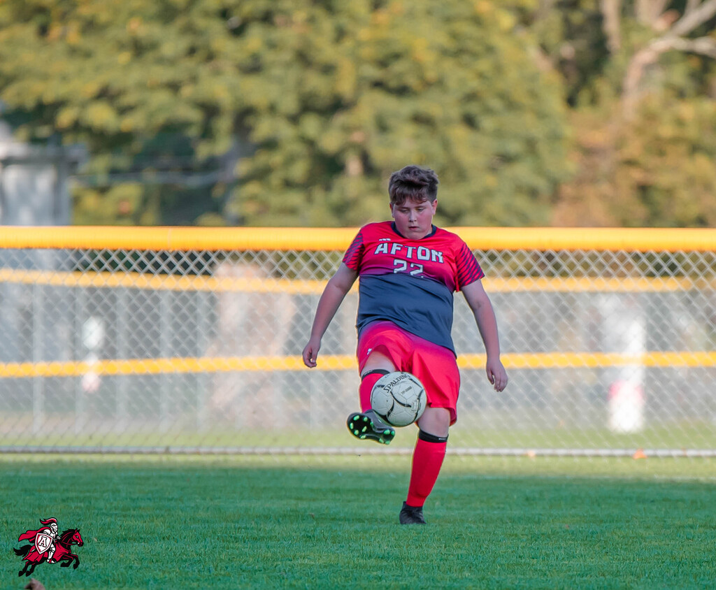 Boy in red and blue soccer uniform, number 22, kicking a ball on a field with a chain-link fence.