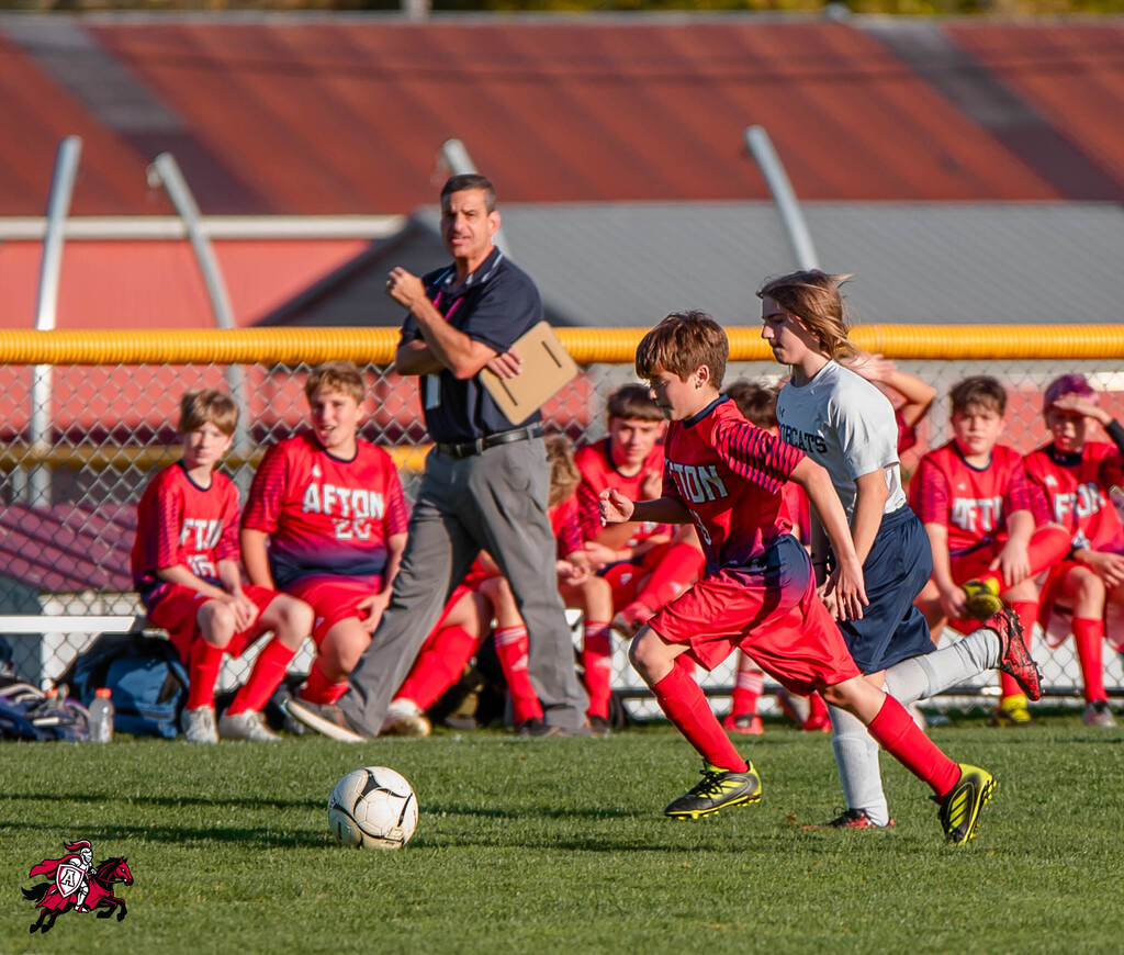 Soccer match with a man in gray pants and a clipboard near children playing. Two children in red and gray uniforms in mid-kick.