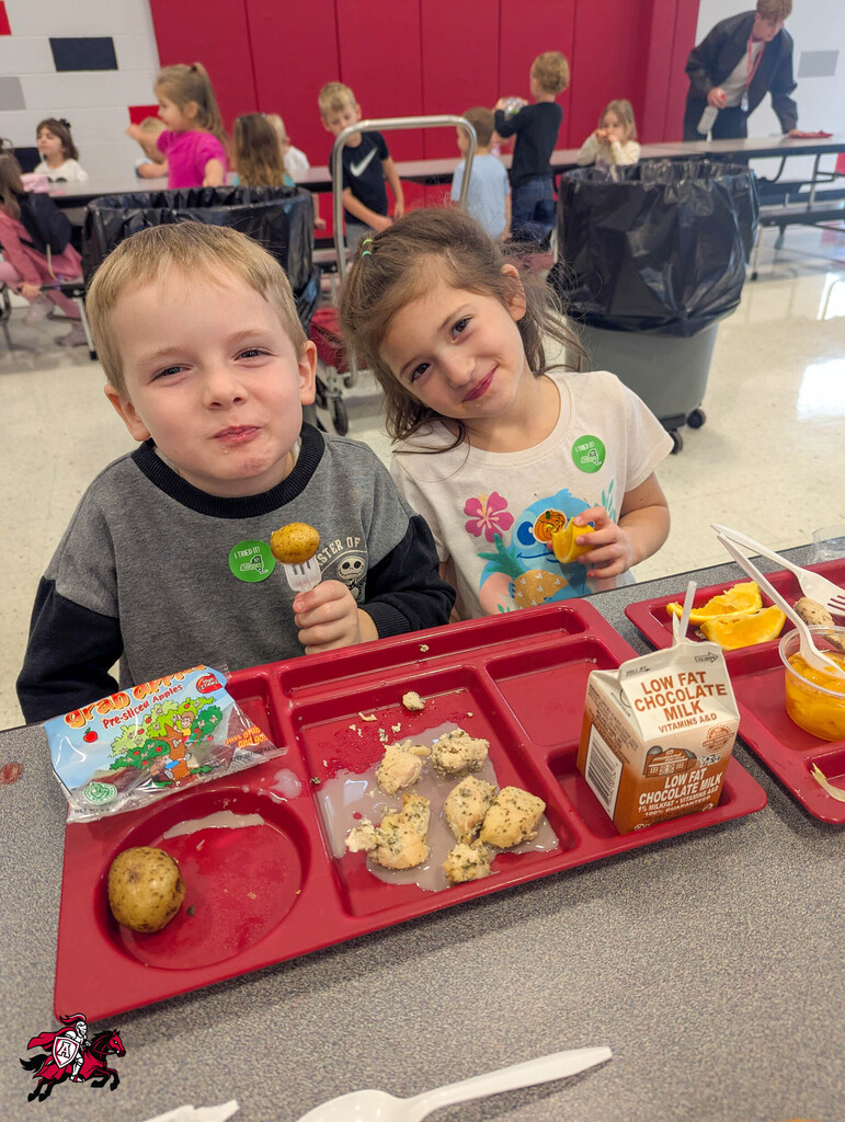 Two children sitting at a lunch table with food trays. A boy has a potato and cookies; a girl has chocolate milk and cookies.