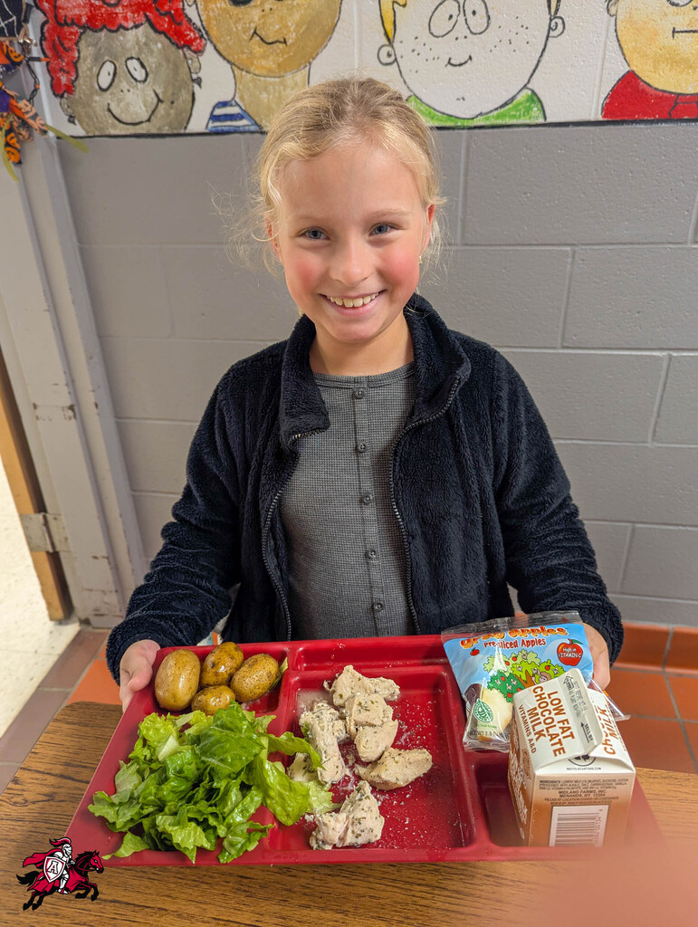 A young girl holds a tray with food, smiling. Tray includes lettuce, chicken, and potatoes. Behind her is a wall with drawings of children.