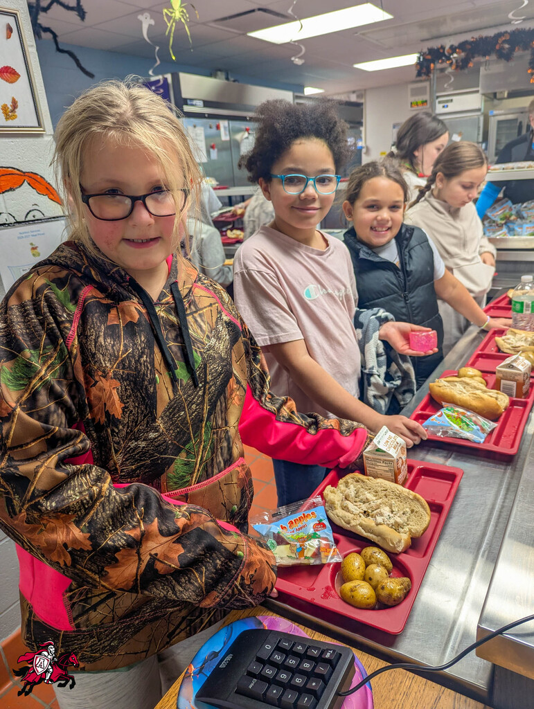 A cafeteria with several children. A girl in camo and glasses is holding a tray of food.