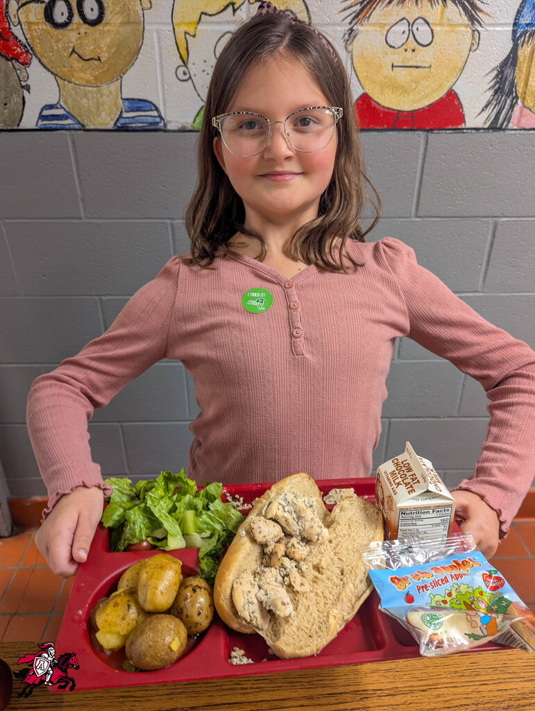 Child in glasses holds a red lunch tray with sandwich, salad, potatoes, carton, and snacks.