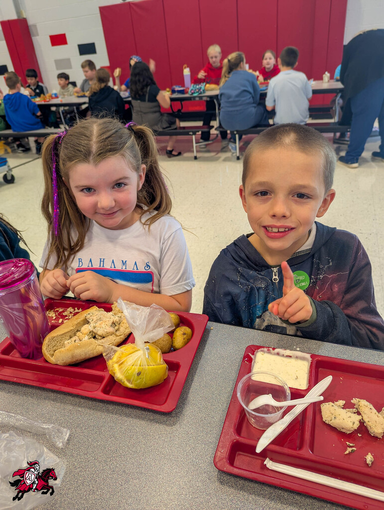 Two children seated at a table, one with a sandwich, the other with a cookie. Red trays hold food and utensils.