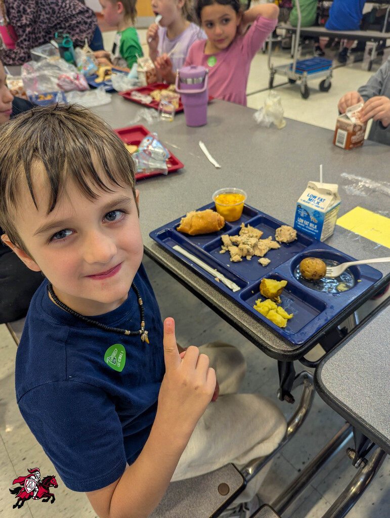 A child sits at a cafeteria table with a meal tray, smiling. Behind him, other children are eating.