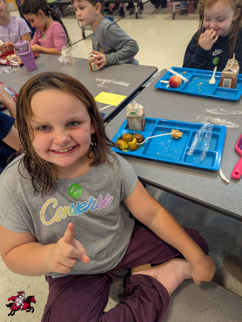 A girl sits at a cafeteria table, smiling and giving a thumbs up. Trays with food, a bottle, and other kids are present.