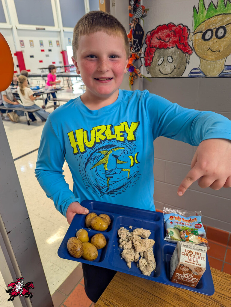A boy holds a cafeteria tray with potatoes, chicken, and juice in a school cafeteria.
