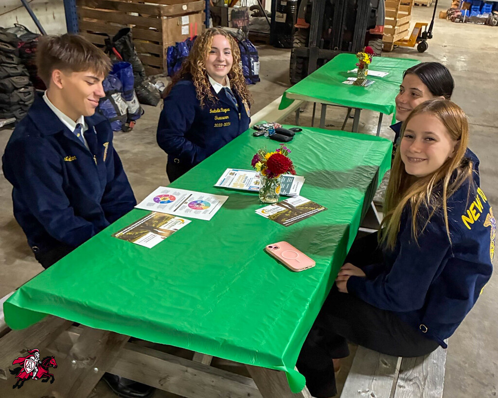 Four students seated around a table covered in green. Papers and flowers on table. One student smiling at the camera.