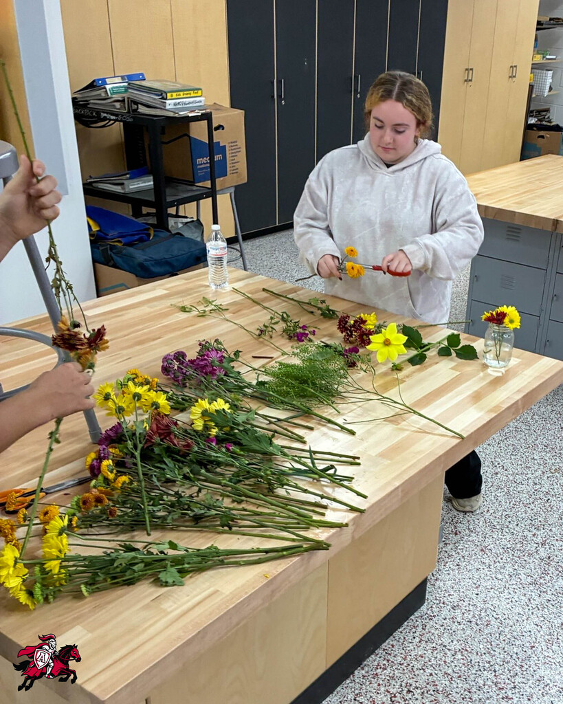 A person with a hoodie and a student with a ponytail arranging flowers on a table.