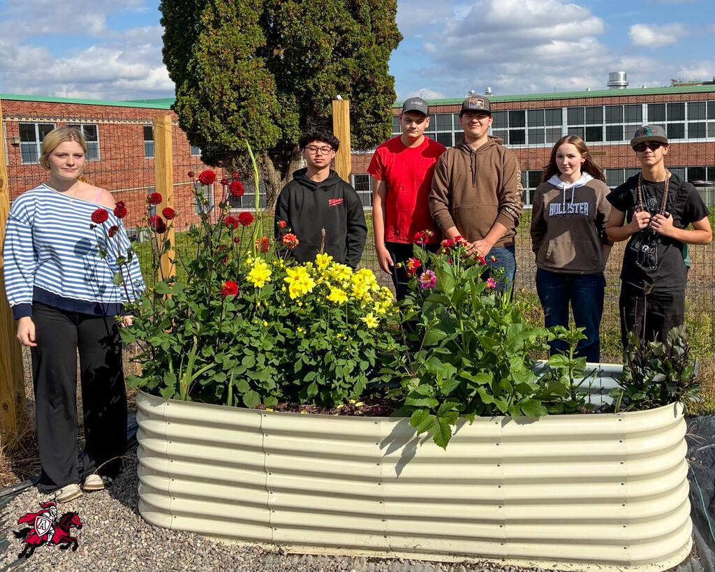 Students and adults posing near a metal planter with flowers and a tree in the background.