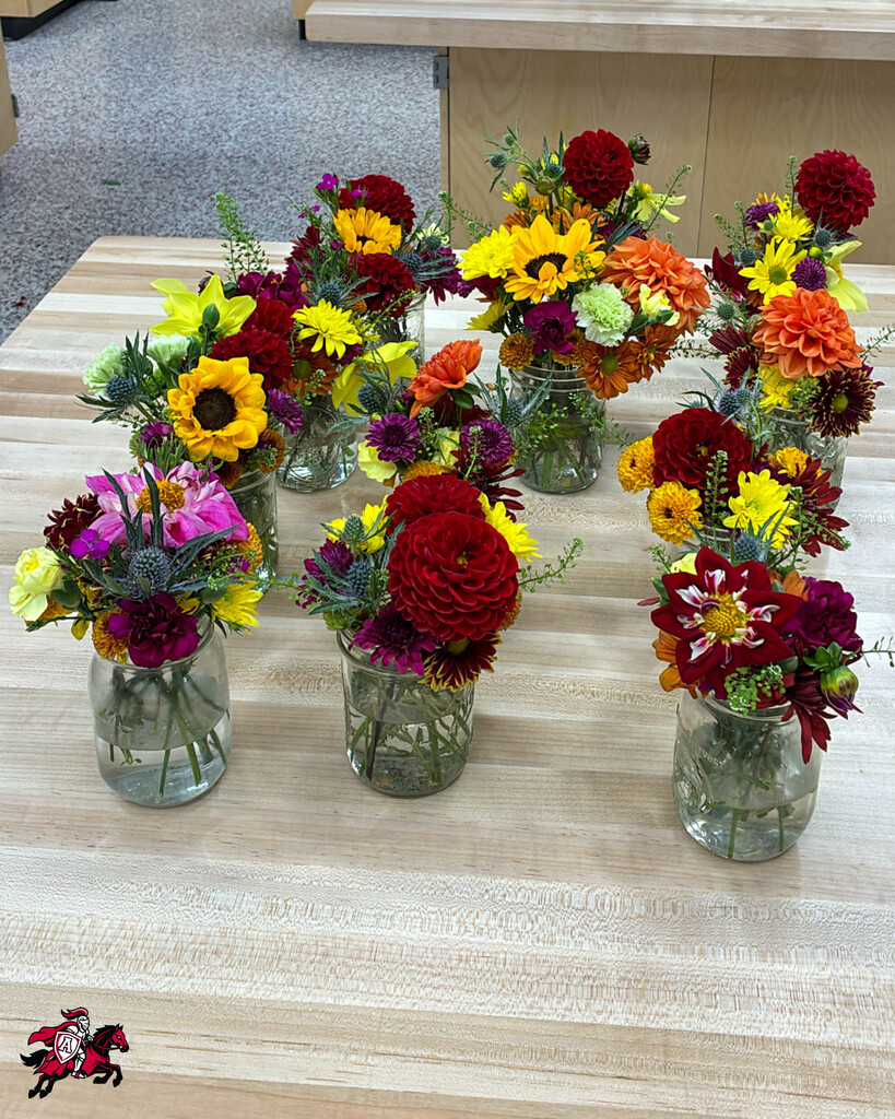 Nine small mason jars with flowers in them on a wooden table with a logo in the lower left corner.