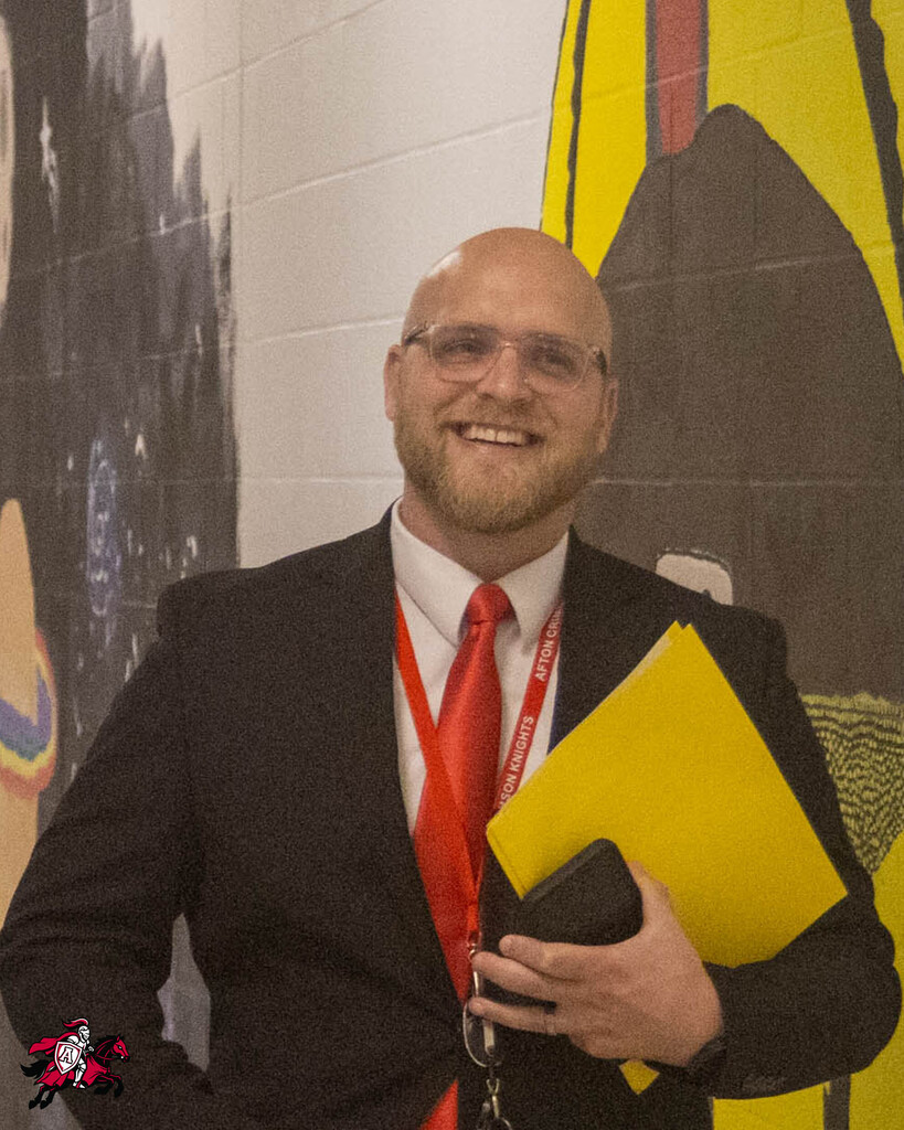 Man in a suit and tie holds a folder and wallet, standing in front of a mural.