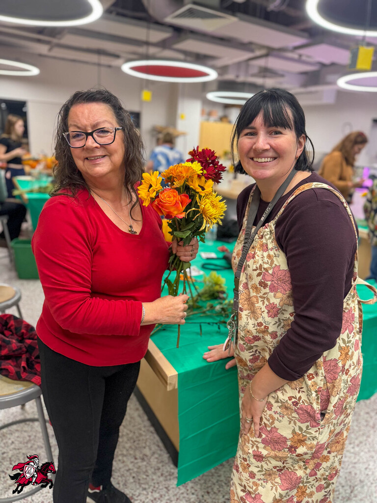 Two women in a room. One holds flowers. Both wear eyeglasses. Behind them, people work.