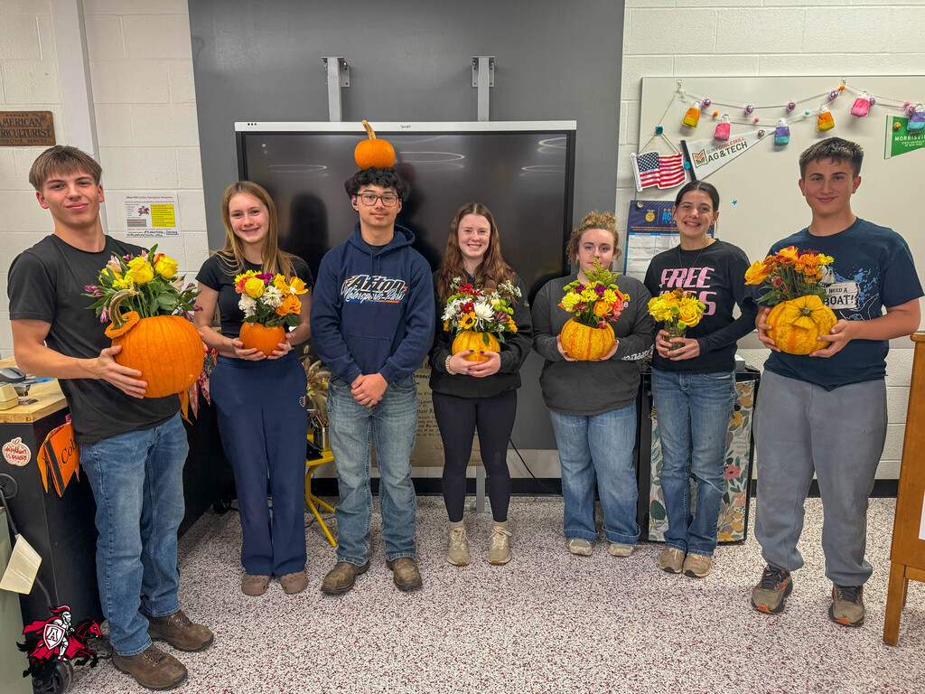 A group of students pose in a room, each holding a small pumpkin with flowers, standing near a whiteboard.