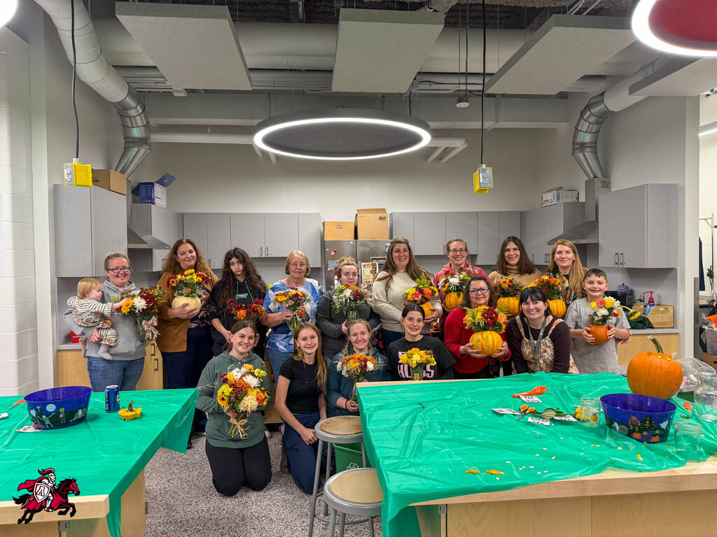 Group of students and adults in a kitchen area, some holding flowers and pumpkins, smiling for a photo.