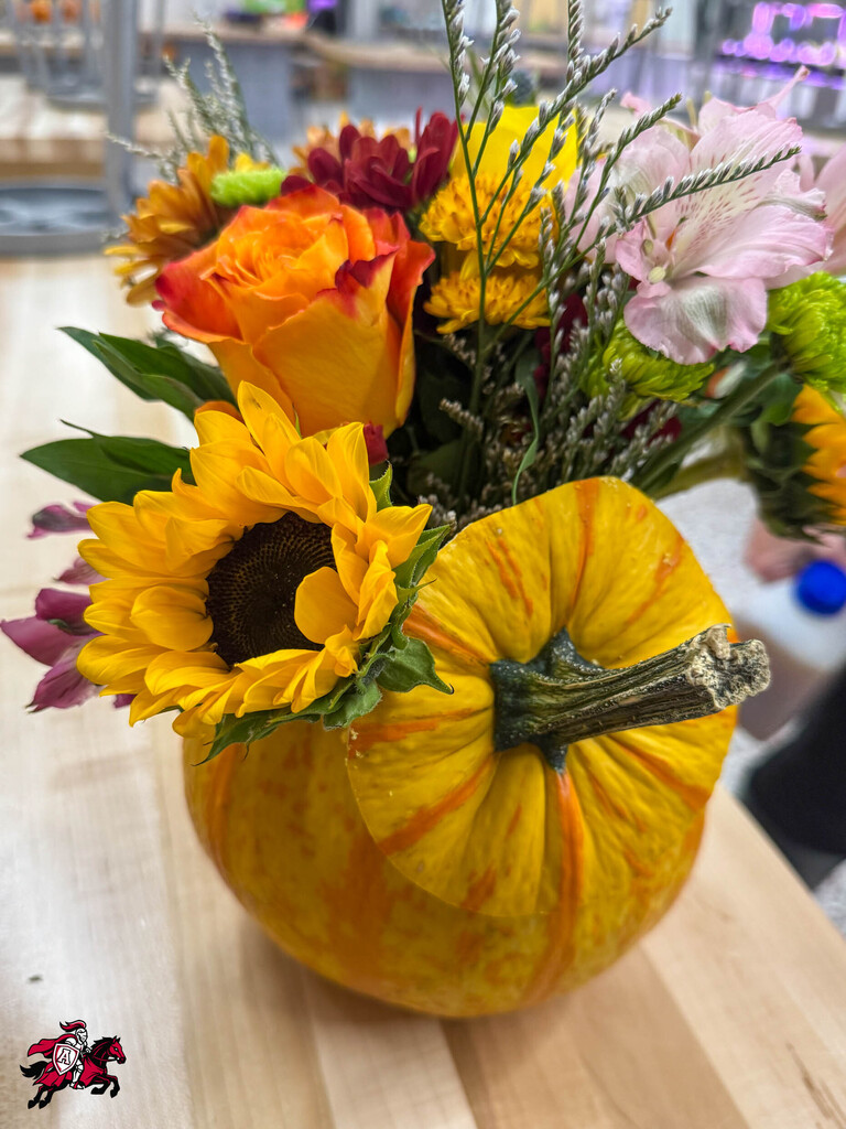Flowers arranged in a pumpkin vase, including sunflowers, roses, and other flowers, on a wooden table.