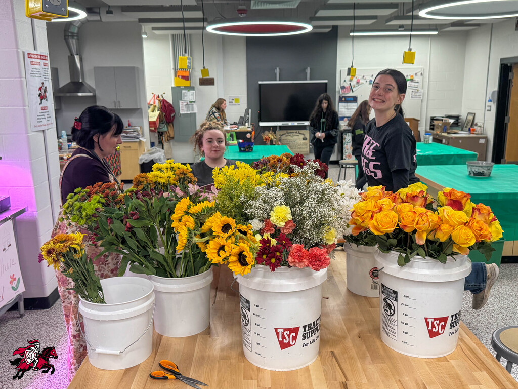 Four students arrange various flowers in buckets on a table in a room with tables and a television.