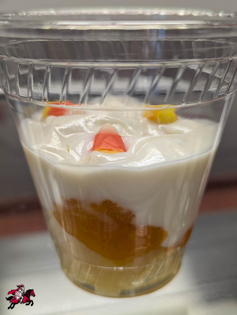 A clear plastic cup with a layered dessert, including a white topping, orange bottom layer, and candies on top.