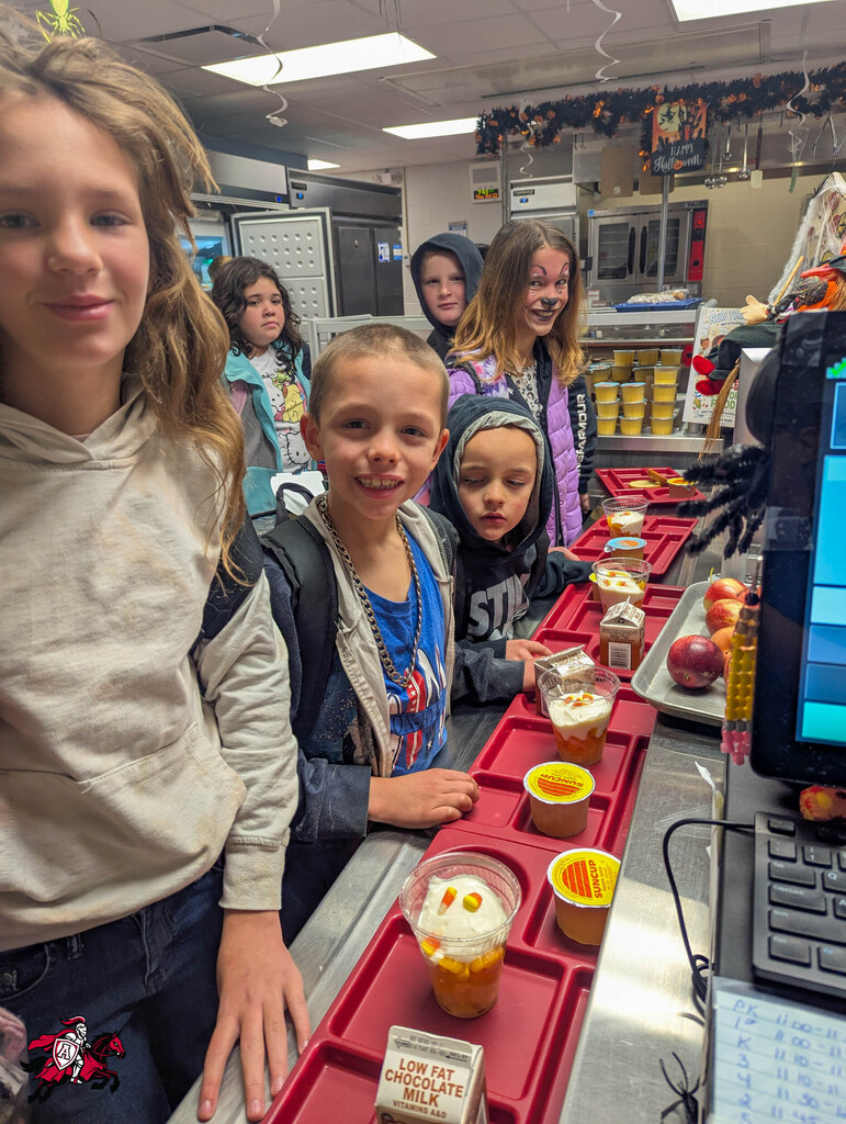 Students stand behind a cafeteria counter, looking at a tray with desserts in cups.