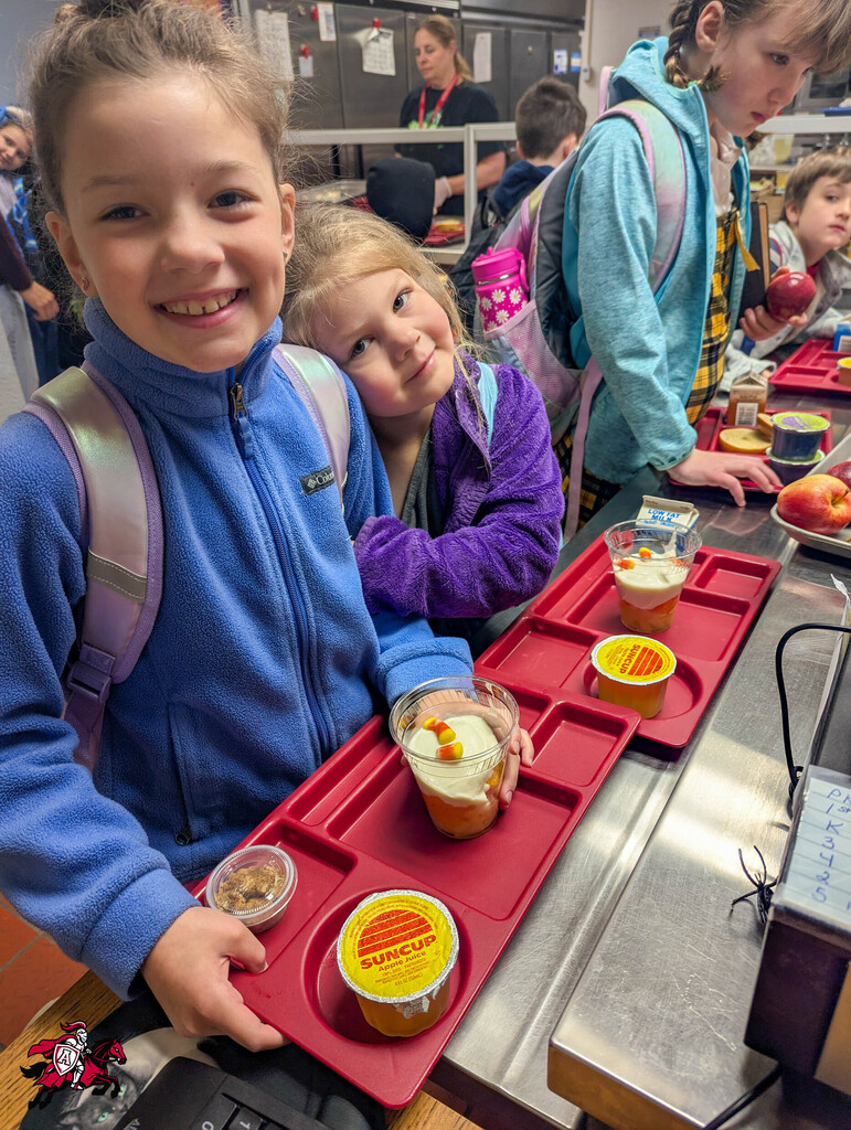Children in blue coats hold trays with food and desserts in a cafeteria with other students.