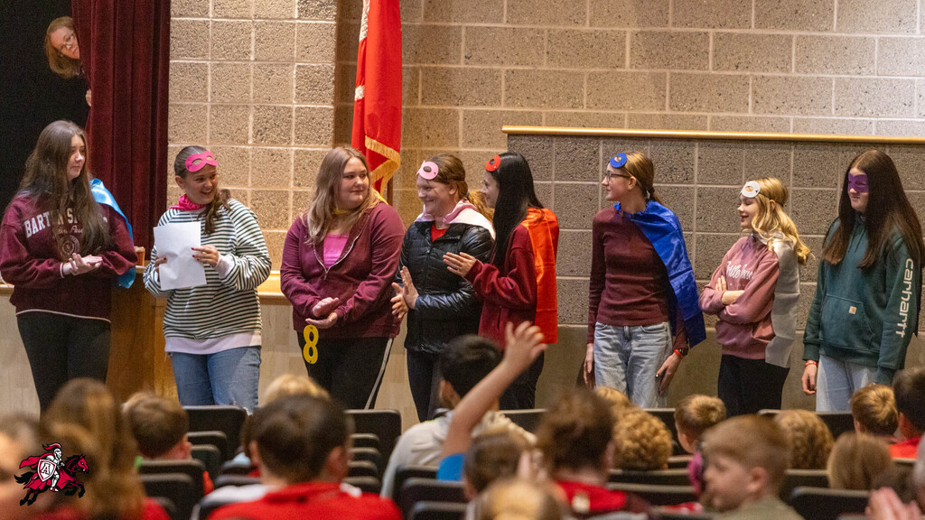 Students standing on stage, some holding papers, while audience sits in front of them in chairs.