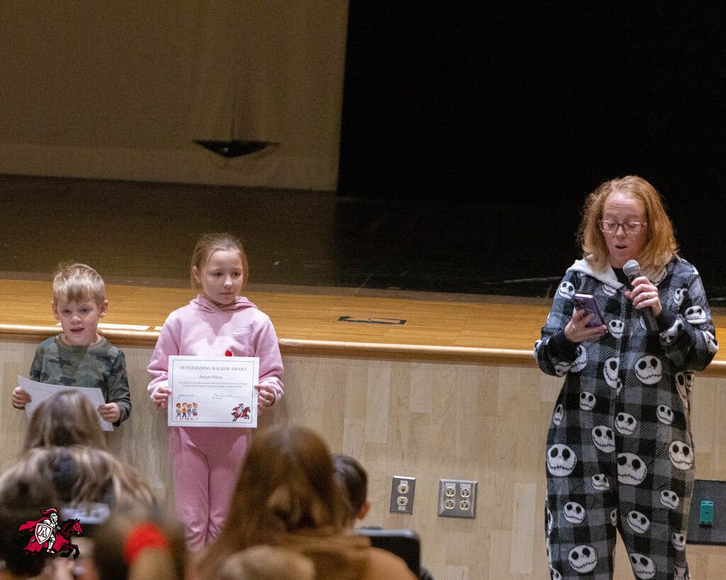A woman with a microphone stands on a stage beside three children holding papers. Behind them is a stage with a curtain and a wooden wall.