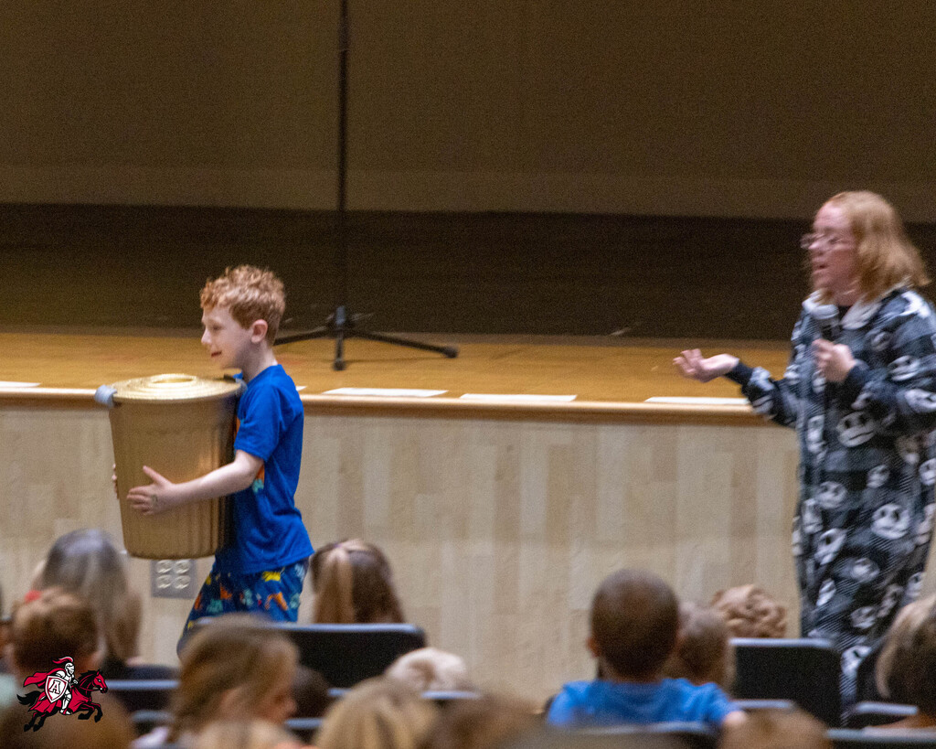 A woman on stage holds a microphone while a young boy with a large object looks at her.