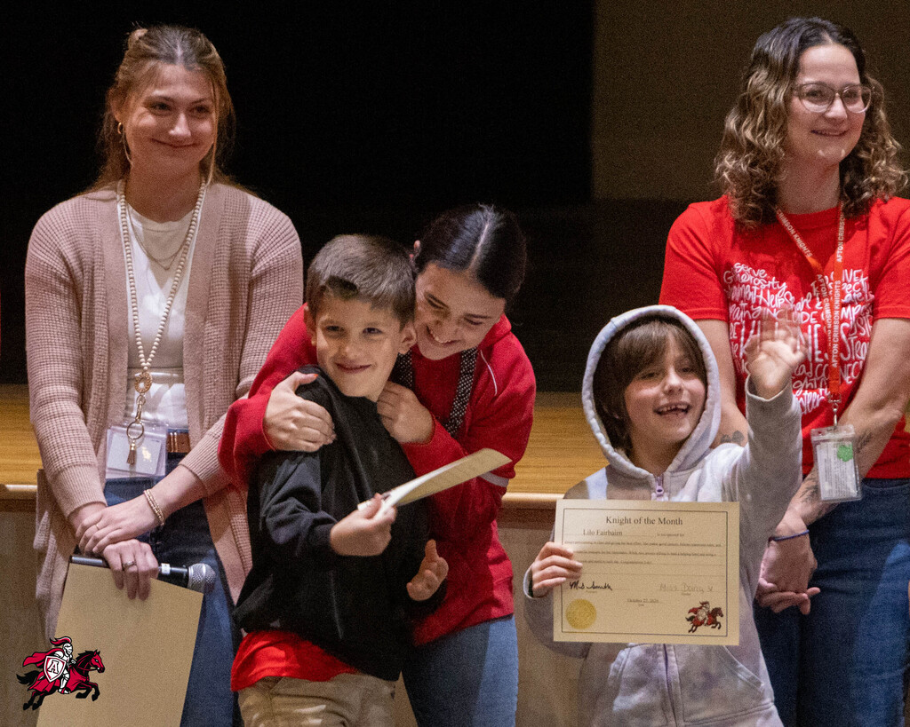 Four adults and two children on stage, smiling and holding certificates. One child raising arm.