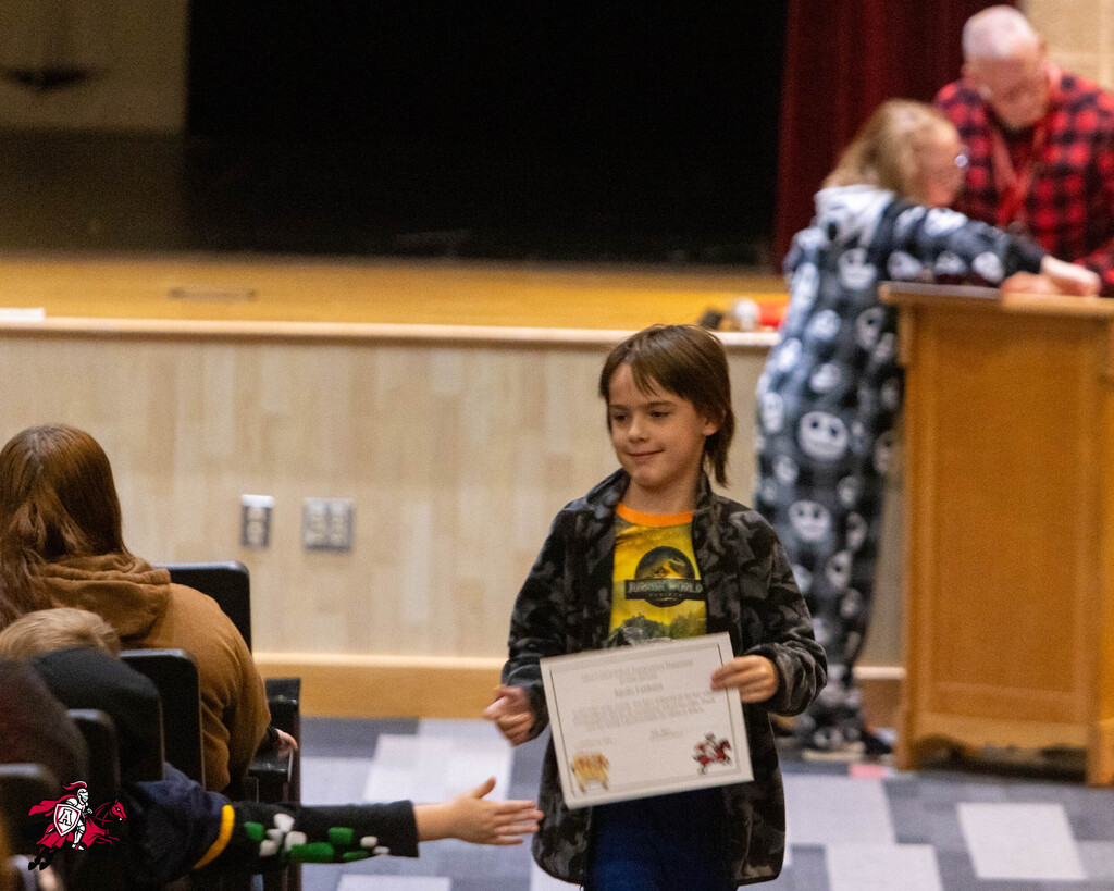 Child holding a certificate stands in front of an audience with a stage and people seated in chairs.