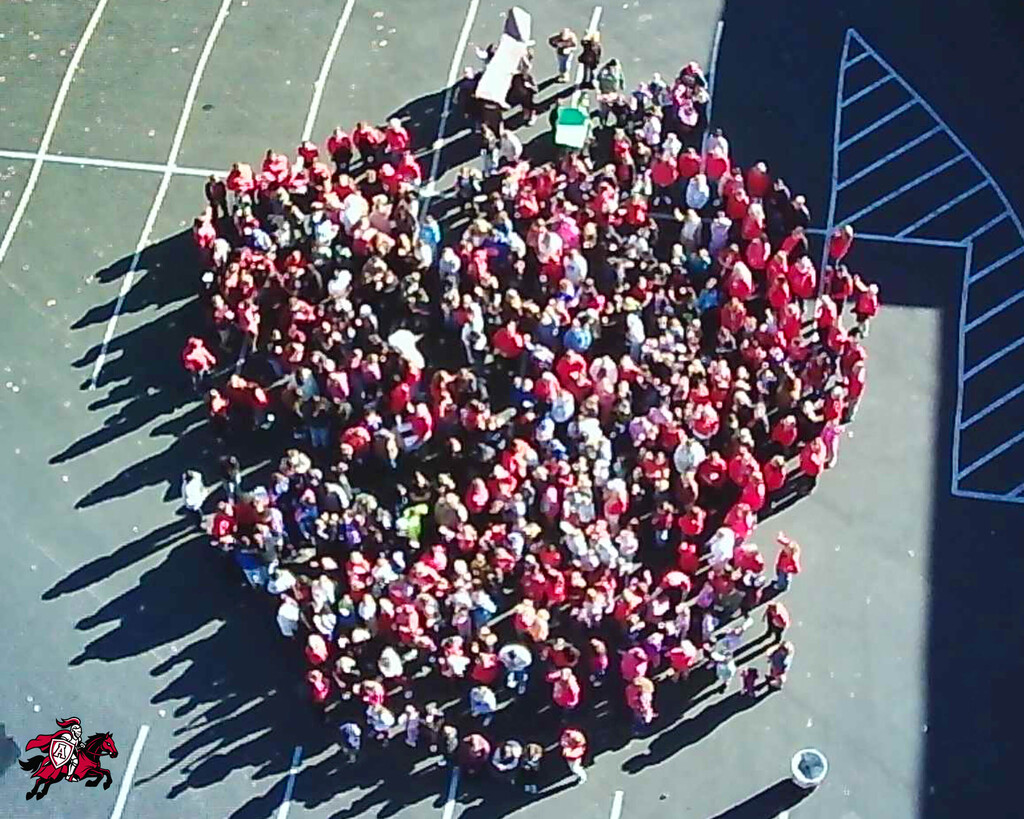 Aerial view of a large group of students in red shirts gathered in a circle on a school's paved parking lot.