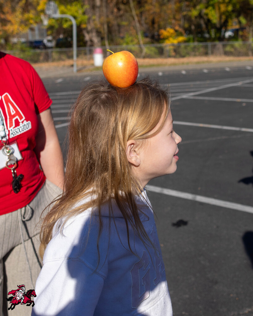 A girl in a white shirt balances an apple on her head while standing on a paved lot.