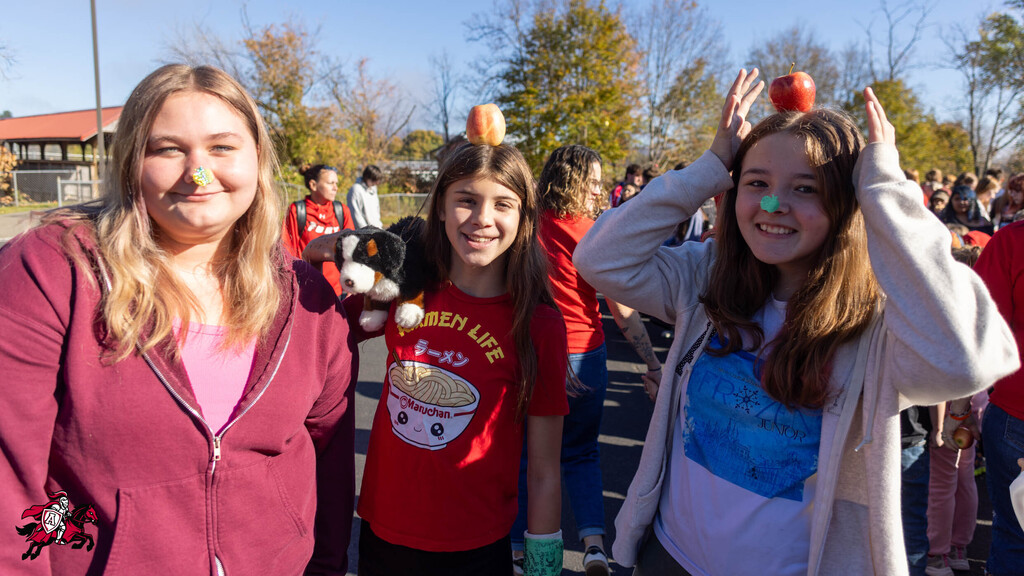 Three girls outside on a day; two have apples on their heads, one holds a stuffed animal.