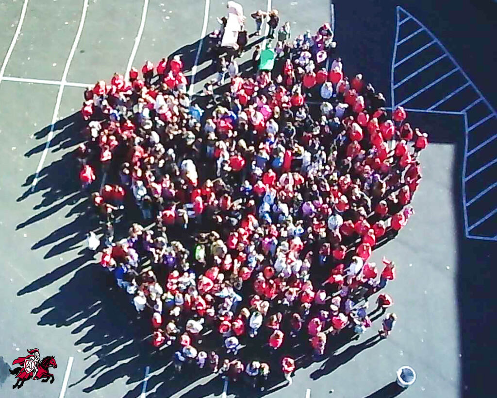 Overhead view of many students in red shirts, gathered in a circle with shadows on a paved surface.