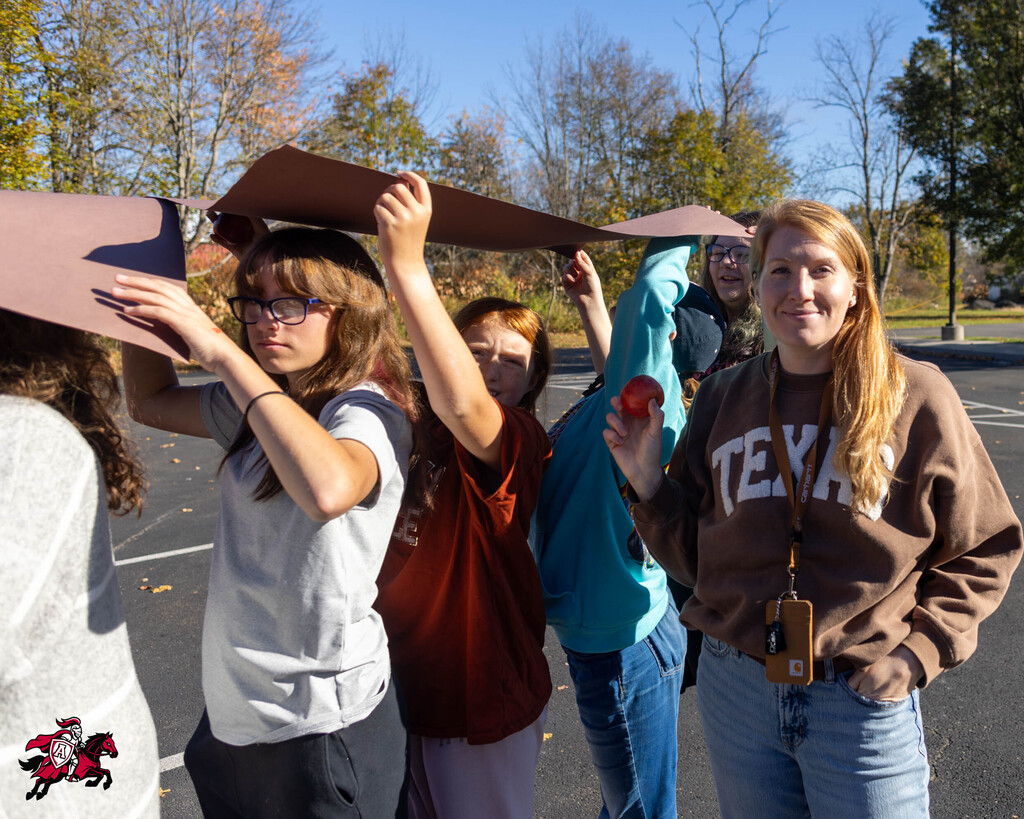 Five students hold paper constructions over their heads in a parking lot; one holds a red object.