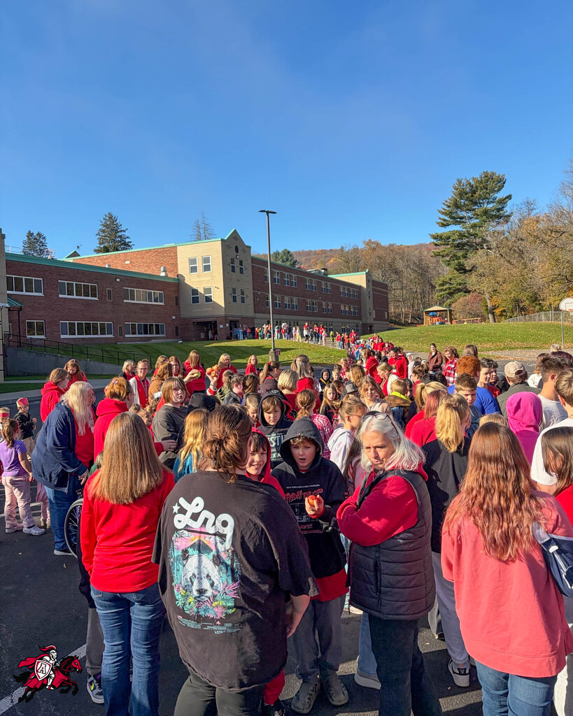 Crowd of people wearing red and black clothing gathered in a parking lot with a large school building in the background.
